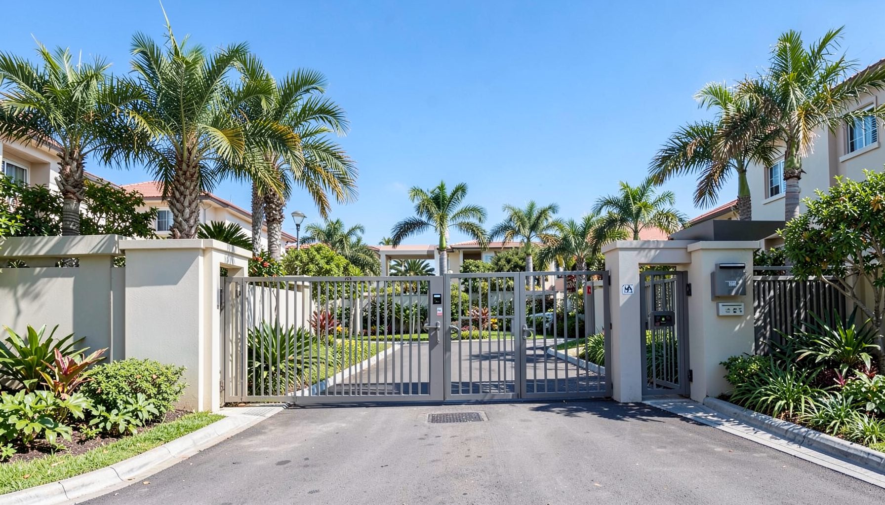Gated residential community entrance with landscaped gardens, palm trees, and modern security gate under a clear blue sky