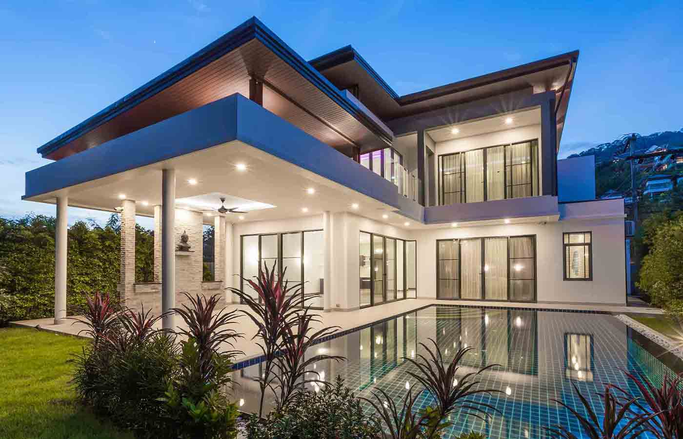 Modern two-story house with large windows, illuminated interior lighting, swimming pool in foreground, tropical plants, warm evening atmosphere