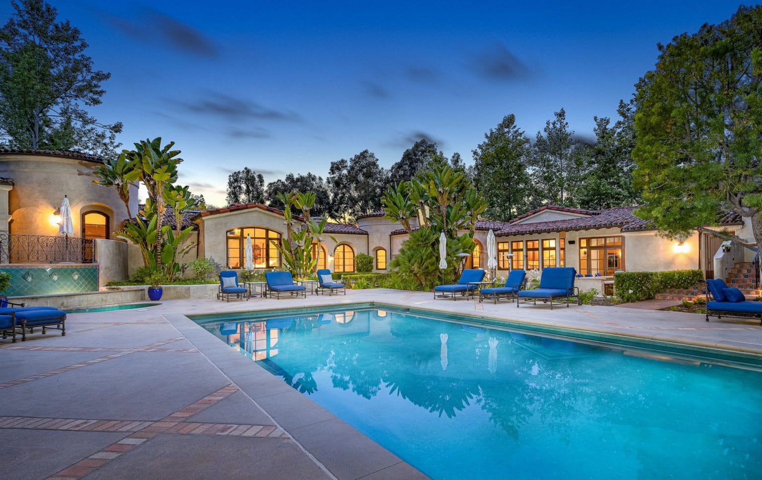 Twilight view of a large Mediterranean-style house with tiled roofs, palm trees, blue lounge chairs and a lit swimming pool, warm and inviting evening mood