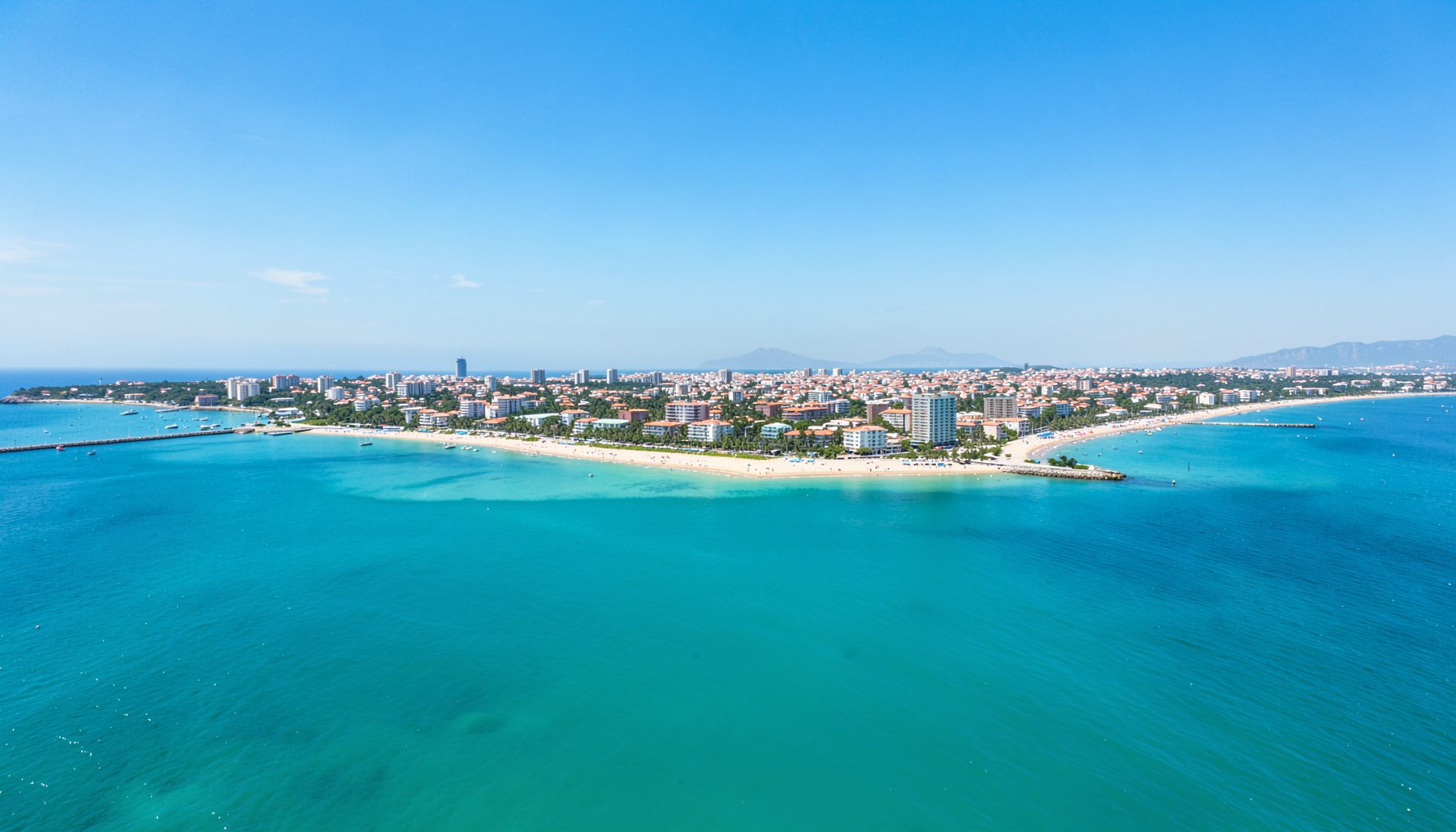 Wide aerial view of a coastal city with turquoise sea, sandy shoreline, and low-rise buildings under a bright blue sky