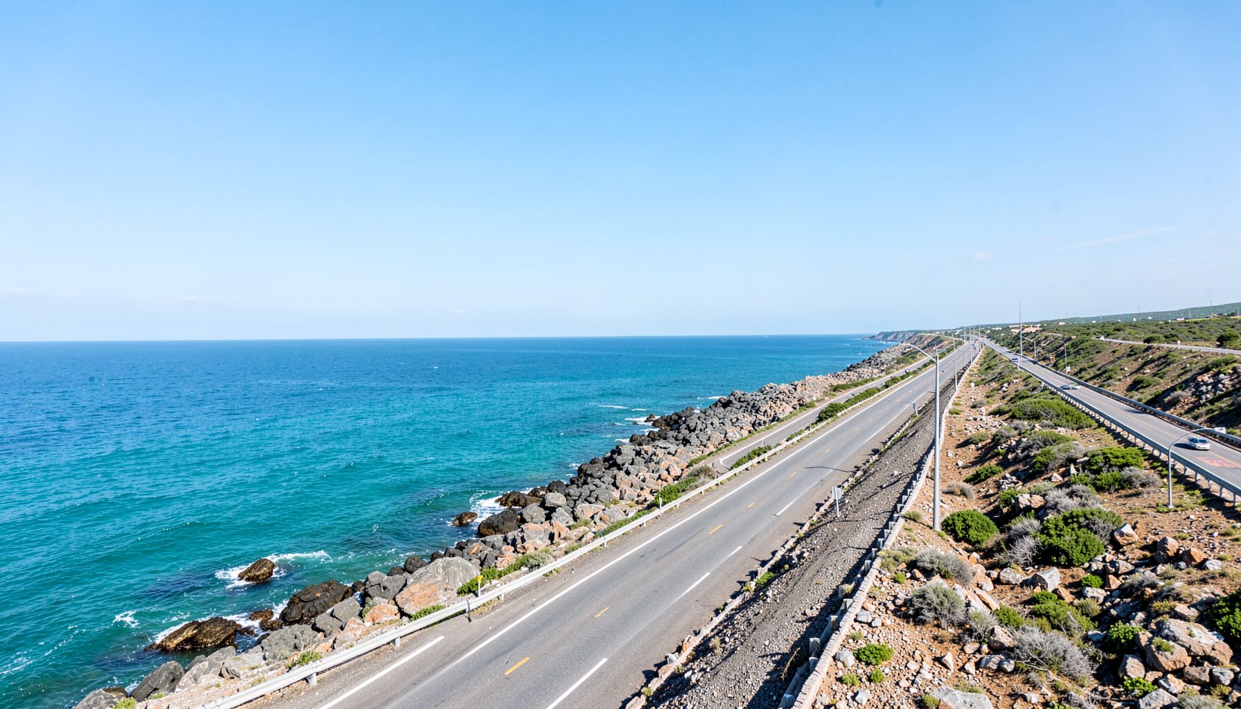 Wide panoramic view of a coastal highway stretching along a rocky shoreline with turquoise ocean under a clear sky
