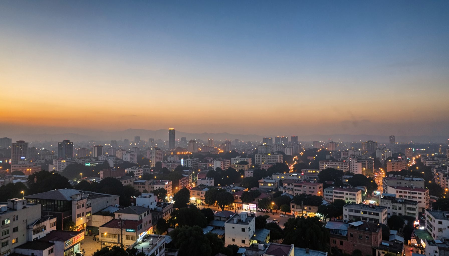Wide view of a modern Lahore cityscape at dusk with warm amber lights, soft haze over distant buildings, and a calm sky